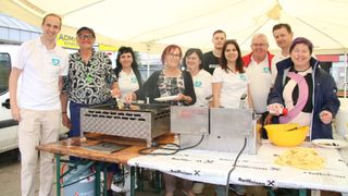 Eine Gruppe von Menschen steht um einen Grill herum und posiert für ein Foto. Sie tragen weiße T-Shirts mit Logos. Eine Frau hält einen Teller mit Essen. Dahinter steht ein weißes Zelt.