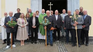 Gruppe von Menschen vor einer Kirche, ein Priester hält ein Kreuz und einen Blumenkranz.