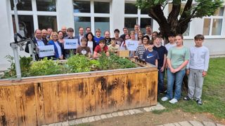 Eine große Gruppe von Menschen steht neben einem Holzpflanzer voller Kräuter und Pflanzen. Einige halten Schilder mit den Aufschriften 'BECOM' und 'MINT'. Sie sind draußen vor einem weißen Gebäude.