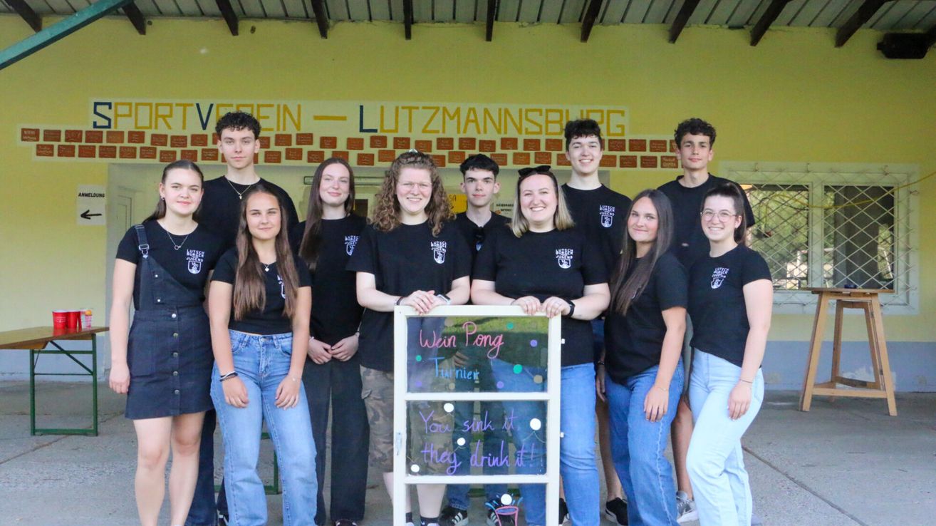 Eine Gruppe junger Menschen in schwarzen T-Shirts posiert für ein Foto vor einer gelben Wand. Ein Schild lautet Wein Pong Turnier.