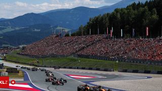 Ein Formel-1-Rennen mit vielen Autos auf der Rennstrecke und einer großen Menge Zuschauer in den Tribünen. Berge im Hintergrund.