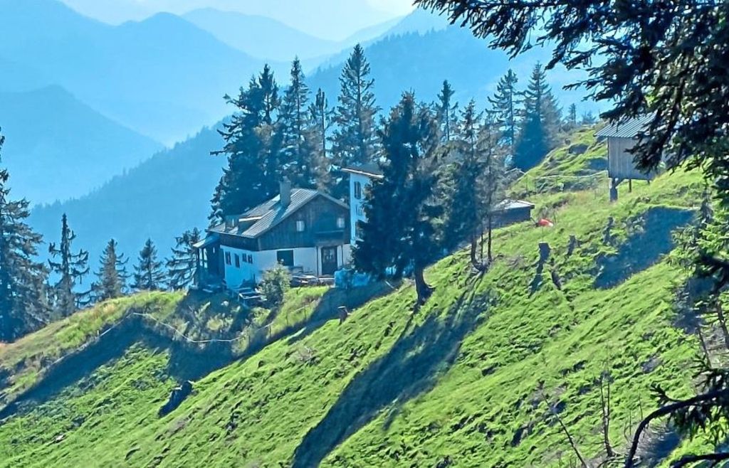 A house with a chimney is nestled on a hillside, surrounded by trees. It overlooks a valley with mountains in the distance.