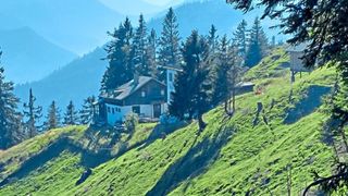 A house with a chimney is nestled on a hillside, surrounded by trees. It overlooks a valley with mountains in the distance.