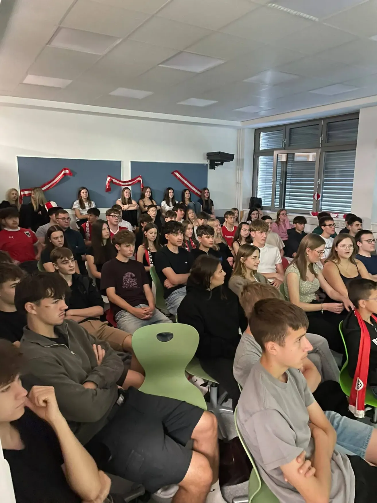 Ein Klassenzimmer gefüllt mit Studenten in lässiger Kleidung. Einige sitzen auf grünen Stühlen, während andere stehen oder auf dem Boden sitzen. Der Raum hat blaue Banner und einen Projektor. Das Glasfenster hat geschlossene Jalousien.
