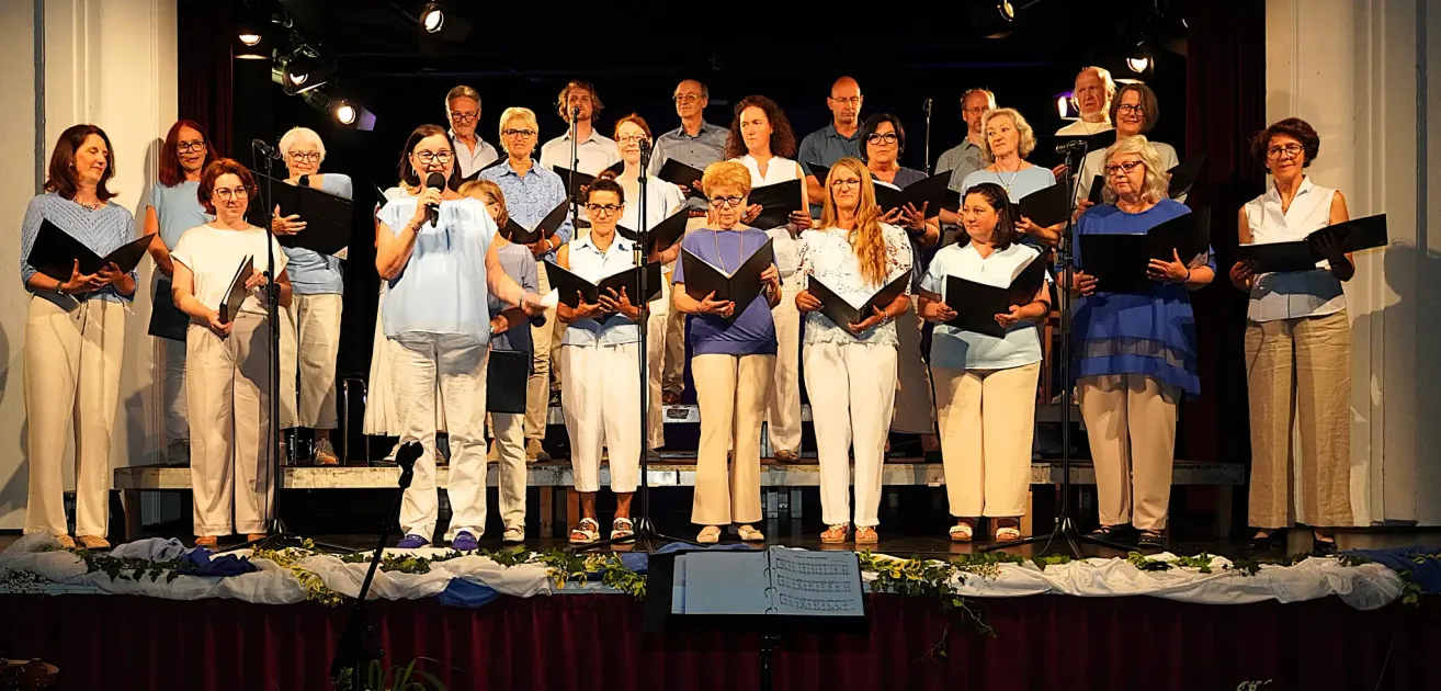 A choir stands on a stage, holding music sheets. The woman in the front is holding a microphone. Behind them, people stand and sing. The stage has a red cloth cover and a music stand.