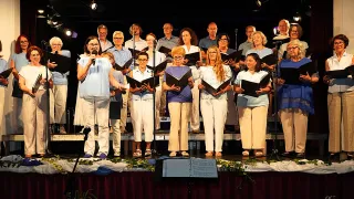 Ein Chor steht auf der Bühne und hält Notenblätter in der Hand. Die Frau vorne hält ein Mikrofon. Hinter ihnen stehen Menschen und singen. Die Bühne hat eine rote Tuchabdeckung und einen Notenständer.