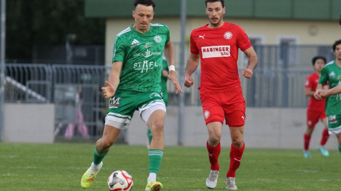 Zwei Fußballspieler auf einem Feld. Einer in Grün, der andere in Rot, beide laufen auf den Ball zu.