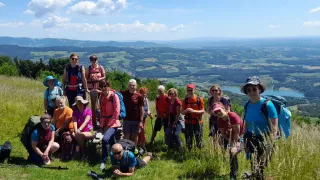 Eine Gruppe von Wanderern, die Mützen und Brillen tragen, posiert auf einem Berg mit Blick auf das Tal. Einige sitzen auf einem Stein, während andere auf dem Gras stehen.