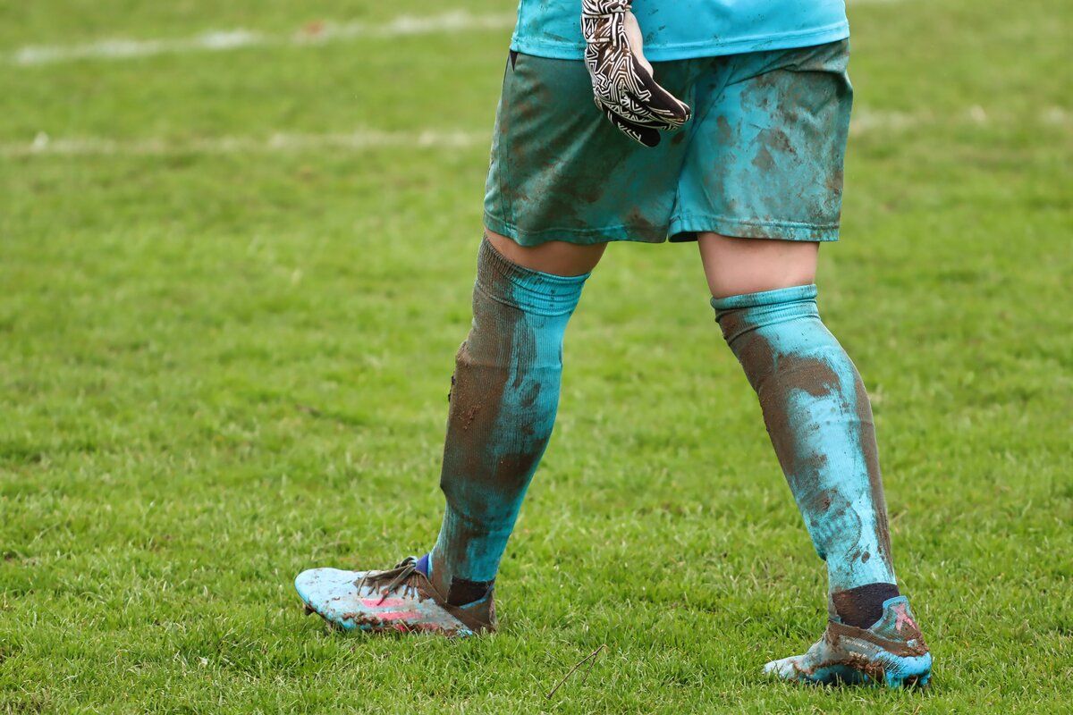 Ein Fußballspieler steht auf einem Feld, die Beine voller Schlamm. Der Spieler trägt blaue Shorts, Socken und Schuhe. Sie halten einen schwarzen Handschuh.