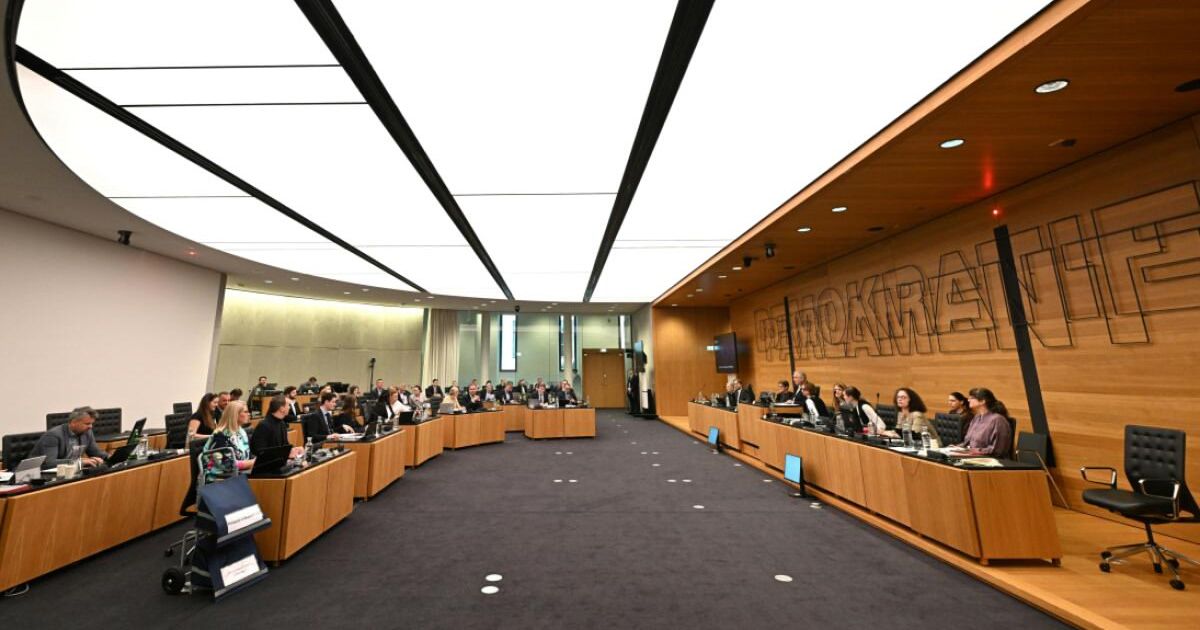 A conference hall with wooden desks, monitors, and participants seated. The walls are wooden with the word 'SPEOKOKA'. The room has a modern design with white ceiling and black carpet.