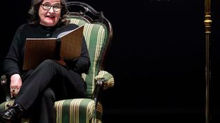 A woman with glasses and a black top sits on an ornate chair, holding an open book and reading.