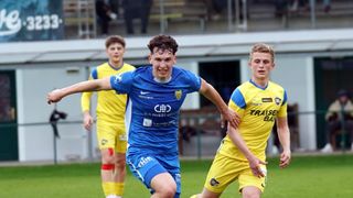 Drei Fußballspieler in blauen und gelben Trikots spielen auf einem Feld. Der Spieler in Blau mit ausgestreckten Armen scheint den Spieler in Gelb zu jagen. Ein anderer Spieler in Gelb steht hinter ihnen und blickt nach hinten.