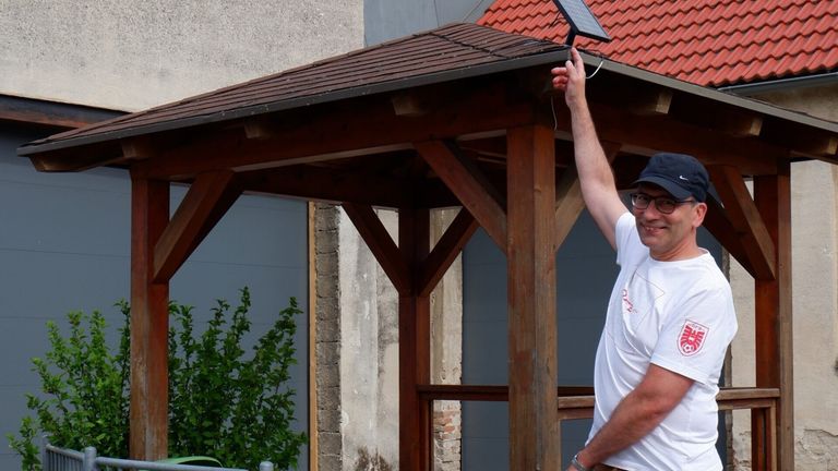 Ein Mann steht unter einem hölzernen Pavillon mit einem Solarpanel auf dem Dach, trägt eine Brille und ein weißes T-Shirt. Links wächst eine Pflanze.