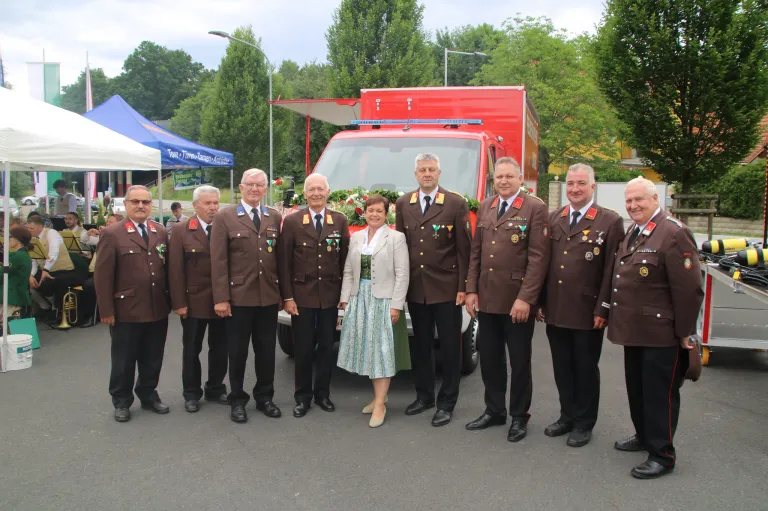 Eine Gruppe von Menschen in Uniform, einschließlich einer Frau, steht vor einem roten Fahrzeug, wahrscheinlich bei einer Veranstaltung. Das Wetter ist sonnig.