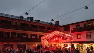 An outdoor performance in front of a building with a crowd watching. The stage is illuminated by red lights, and there are speakers and a microphone on the stage.