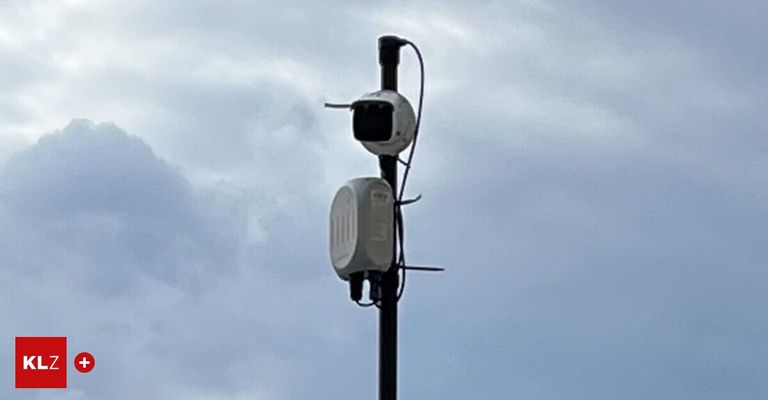 Zwei weiße Geräte sind an einem Mast angebracht gegen einen bewölkten Himmel. Ein Gerät hat eine Kamera oben.