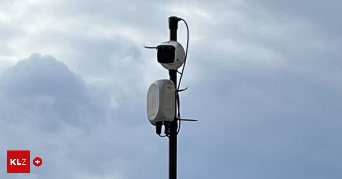 Zwei weiße Geräte sind an einem Mast angebracht gegen einen bewölkten Himmel. Ein Gerät hat eine Kamera oben.