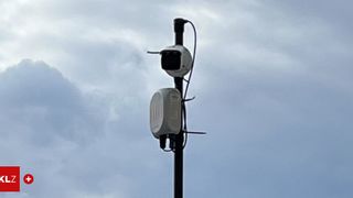 Zwei weiße Geräte sind an einem Mast angebracht gegen einen bewölkten Himmel. Ein Gerät hat eine Kamera oben.
