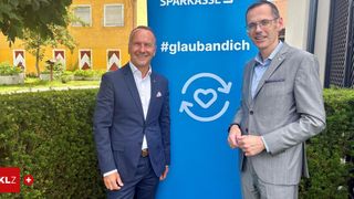 Two men in suits stand in front of a blue sign with the hashtag #glaubandich and a heart symbol. Behind them is a yellow building with red shutters.
