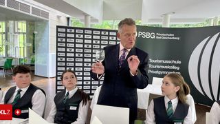 Ein Mann in Anzug und Krawatte hält ein Weinglas und spricht in einem Raum mit zwei Mädchen in Uniform. Hinter ihm steht ein Schild mit Text und Logos.