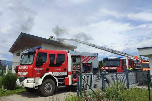 Feuerwehrfahrzeuge sind vor einem Gebäude geparkt, aus dem Rauch aufsteigt. Zwei Feuerwehrleute beobachten das Gebäude, während ein weiterer auf einer Leiter steht.