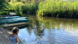 Ein kleines Mädchen steht in einem Fluss und blickt auf das Wasser. Zwei Boote liegen am Ufer festgemacht.