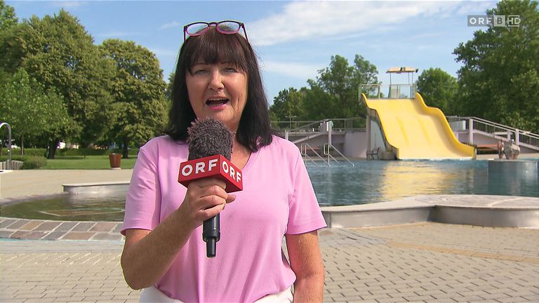 Eine Frau in einem rosa T-Shirt hält ein Mikrofon mit der Aufschrift 'ORF' in der Hand, steht neben einem Pool mit einem gelben Wasserrutsche im Hintergrund.