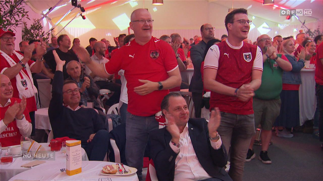Eine Gruppe von Menschen, die rote Trikots tragen und applaudieren, einer von ihnen ist ein Mann in einem Anzug, der auf einem Stuhl sitzt und lächelt, es gibt eine Schachtel und einen Teller auf einem Tisch vor ihnen.