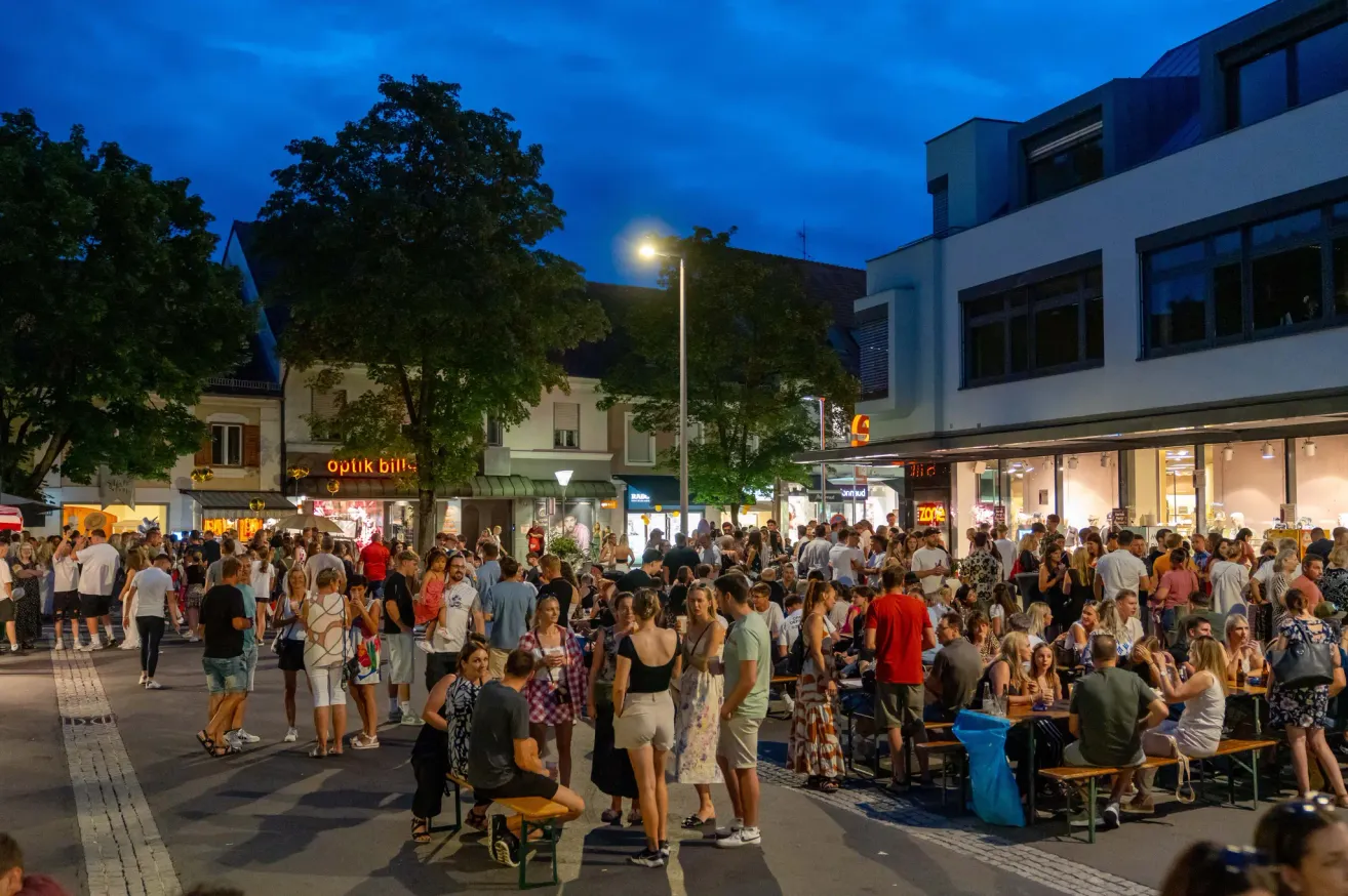 Eine geschäftige Straße bei Nacht mit Menschen, die Getränke an Tischen im Freien genießen und stehend herumstehen, einige unter einem Baum. Straßenlaternen und Geschäfte säumen die Straße.