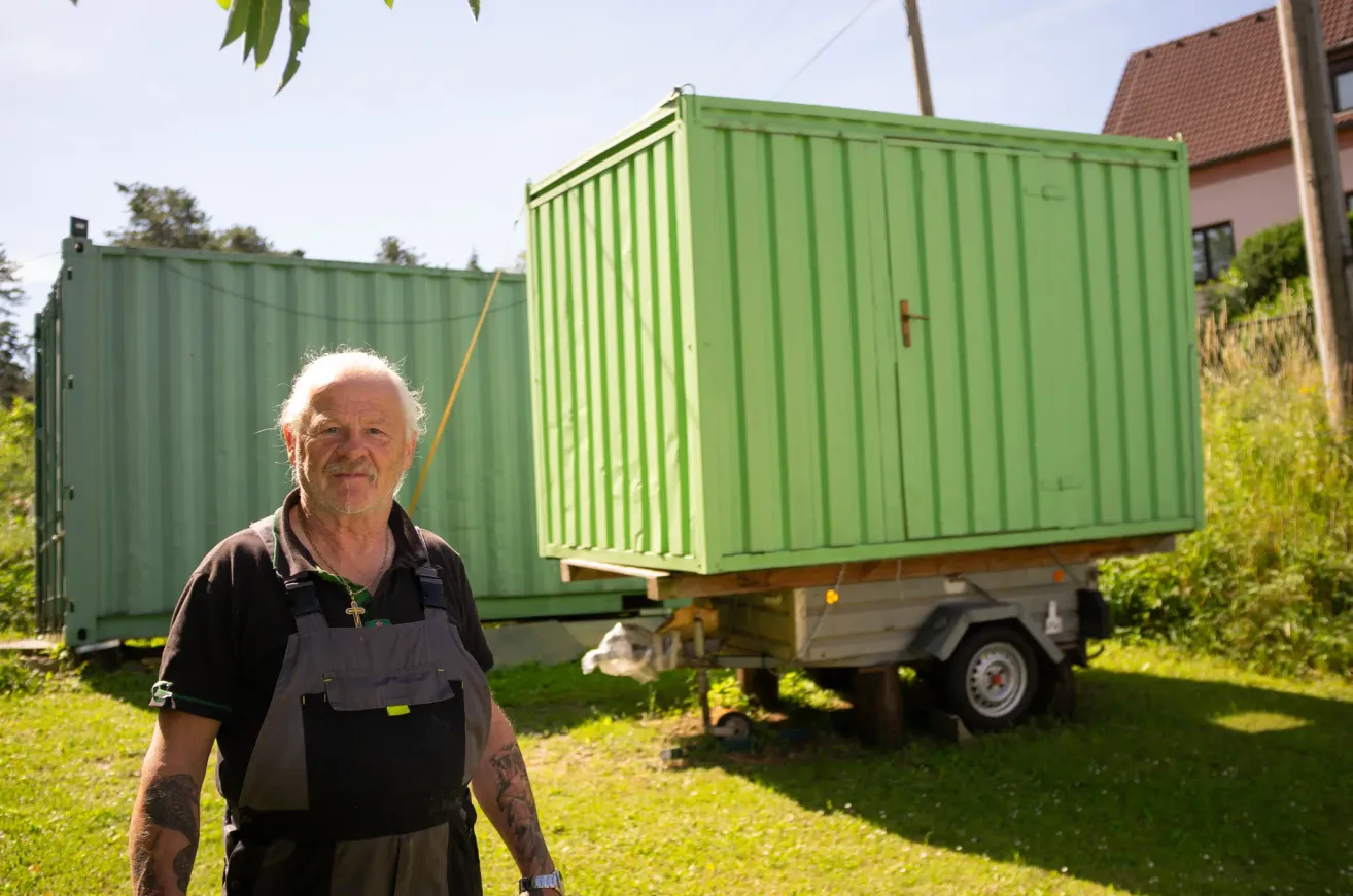 Ein älterer Mann steht vor einem grünen Container, trägt ein schwarzes Hemd und eine graue Latzhose. Der Container steht auf einem Anhänger mit einer Felge und ist von Gras umgeben.