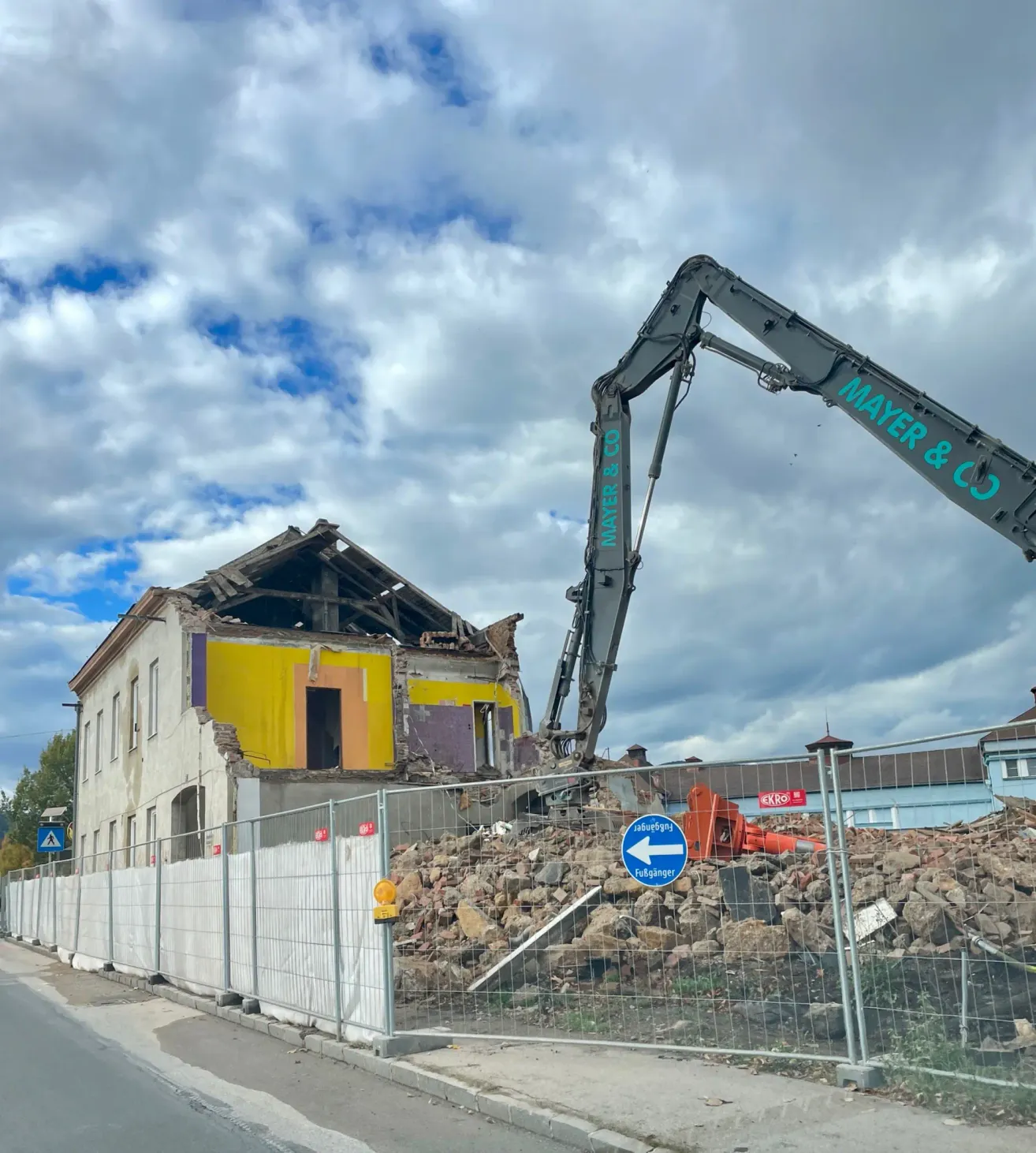Ein Abrissbaustelle zeigt ein Haus in Trümmern, mit einem großen Kran von Mayer & Co. in der Nähe. Schutt liegt auf dem Boden, und ein Zaun umgibt die Baustelle.