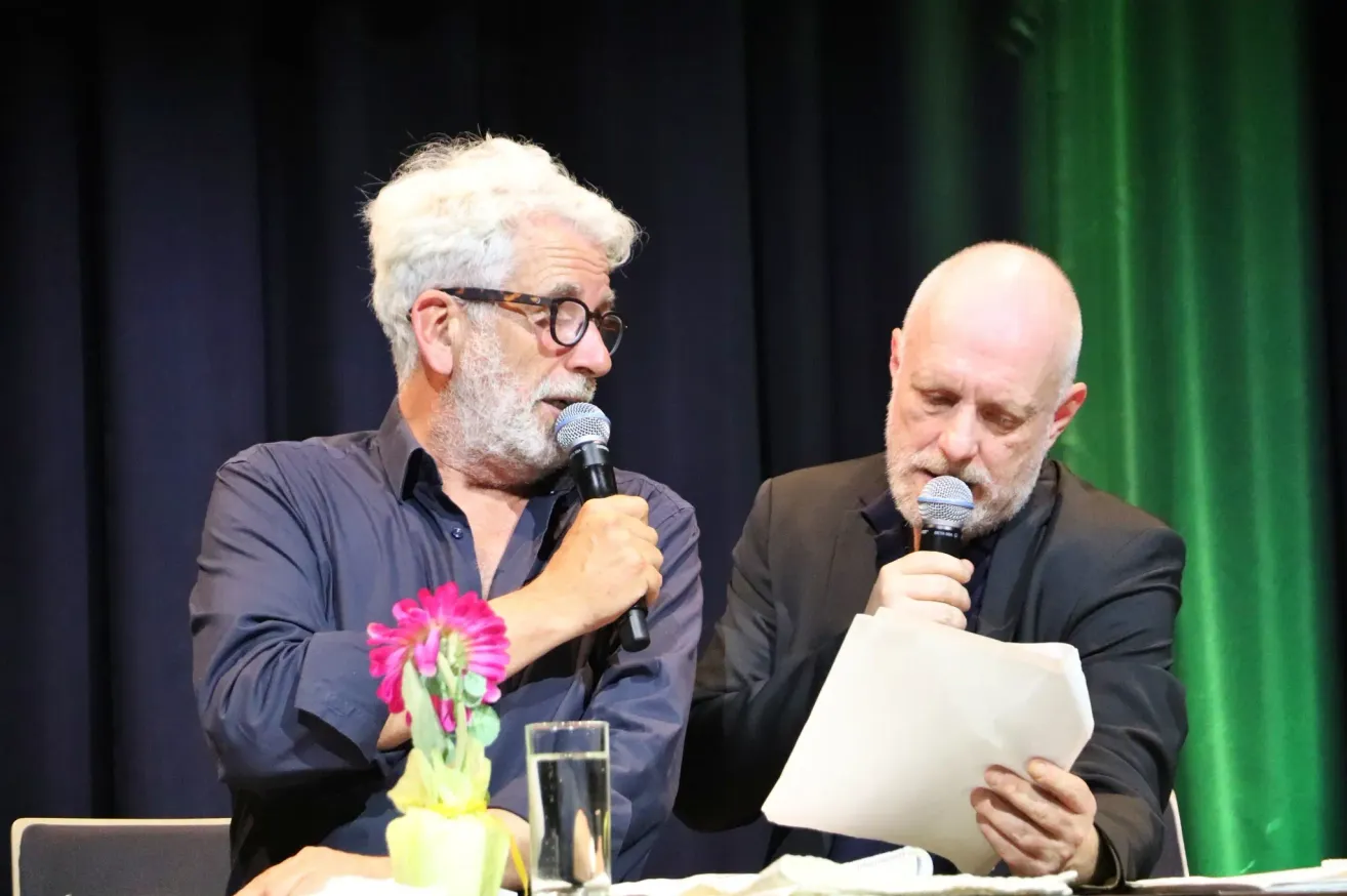 Zwei Männer sitzen mit Mikrofonen an einem Tisch. Der Mann auf der linken Seite hält eine Blume und trägt eine Brille, während der Mann auf der rechten Seite ein Papier in der Hand hält und ernst aussieht.