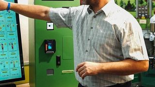 Ein Mann mit Bart steht neben einer grünen Maschine und zeigt auf einen Bildschirm.