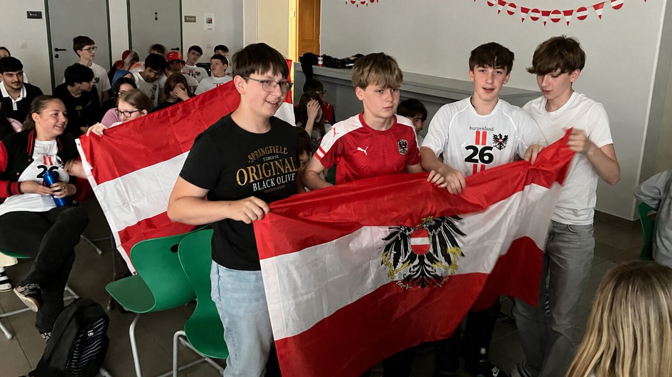 Drei Jungen halten eine große österreichische Flagge, einer trägt ein Springfield Original Black Olive T-Shirt. Dahinter beobachten mehrere andere, einige tragen rote Mützen. Im Hintergrund befindet sich eine weiße Wand mit einer Tür und Dekorationen.