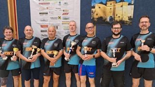 Fünf Männer stehen zusammen und halten Trophäen, wahrscheinlich um einen Sieg zu feiern. Sie tragen passende blaue und schwarze Uniformen. Hinter ihnen zeigt eine Wand verschiedene Logos und ein Bild einer Burg.