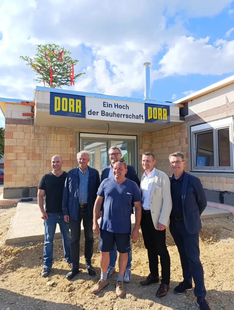 Six men stand in front of a building, smiling. The building has a sign reading 'PORR Ein Hoch der Bauherrenschaft'. Behind them, a tree with red ribbons adorns the roof. The sky is partly cloudy.