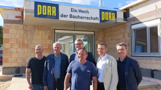 Six men stand in front of a building, smiling. The building has a sign reading 'PORR Ein Hoch der Bauherrenschaft'. Behind them, a tree with red ribbons adorns the roof. The sky is partly cloudy.