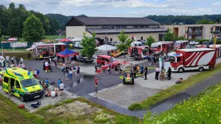 Eine große Veranstaltung mit vielen Menschen, Zelten, Fahrzeugen und Feuerwehrautos. Einige Leute gehen, während andere auf Bänken sitzen. Das Gebiet ist von Gras und Bäumen umgeben.