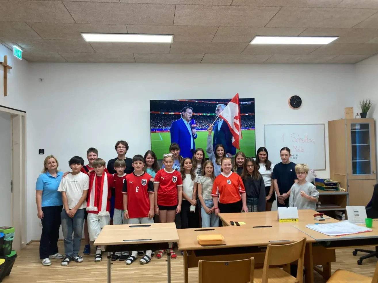 Eine Gruppe von Schülern steht vor einem Klassenzimmer mit einer großen Leinwand, auf der zwei Männer zu sehen sind. Sie tragen rote Trikots mit Nummern und einige halten eine Flagge. Ein Whiteboard befindet sich hinter ihnen.