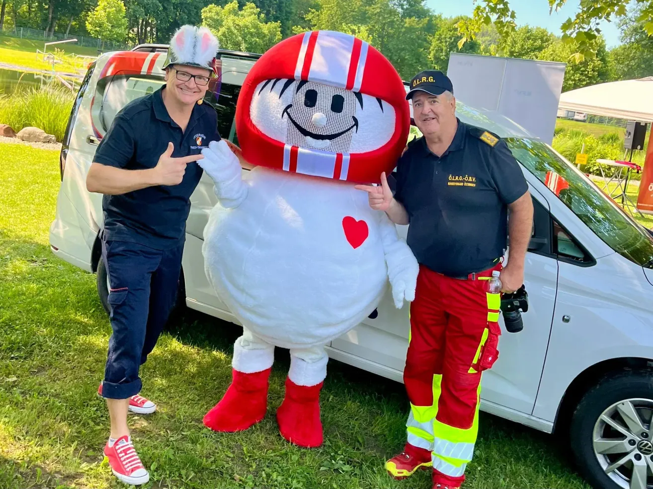 Two men in uniforms, one with a bunny hat, pose next to a mascot and a white van. Trees and a tent are in the background.