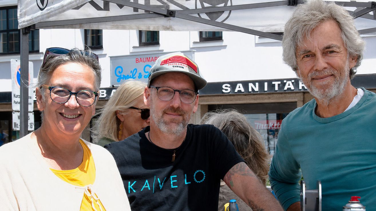 Ein Mann mit einem KaveLo-Shirt und einem Hut lächelt für die Kamera mit zwei anderen Leuten, vor einem Gebäude mit einem Schild, das 'Sanität' steht.