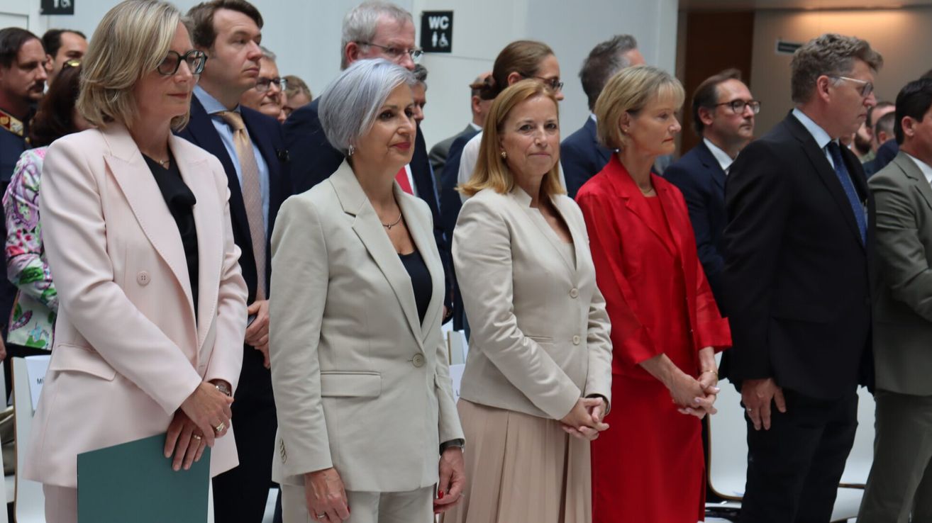 Mehrere formell gekleidete Personen stehen in einem Raum, möglicherweise bei einer Veranstaltung. Zwei Frauen in Anzügen sind prominent zu sehen.