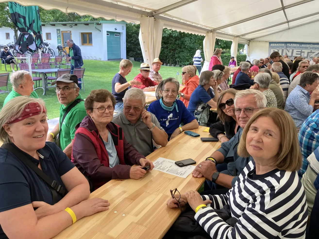 A group of older people are sitting at a long wooden table in a tent, with others around them. There is a building and trees in the background.
