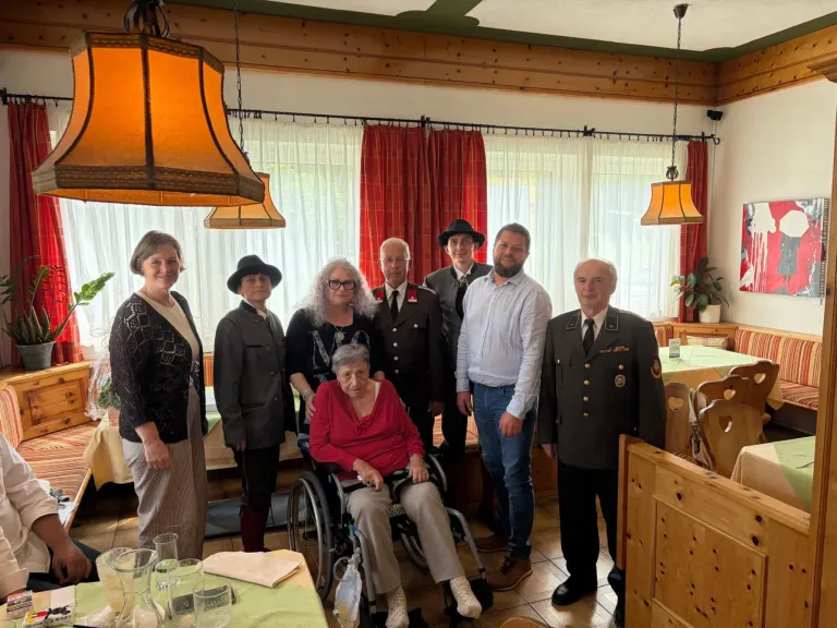 Eine Gruppe von Menschen in einem Raum mit einer Frau im Rollstuhl, die in formeller Kleidung gekleidet ist, stehen um sie herum, einige tragen Hüte. Es gibt Vorhänge, Lampen und einen Tisch mit Gläsern.