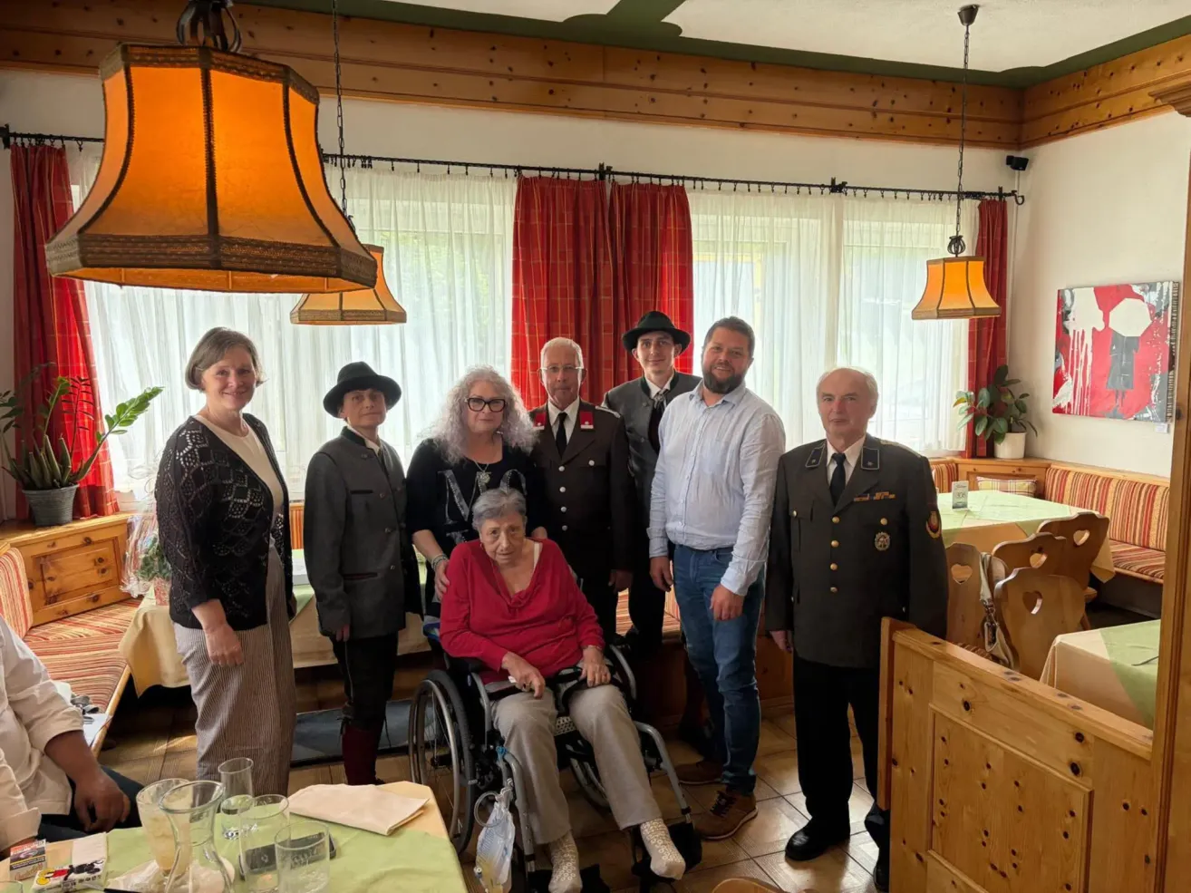 Eine Gruppe von Menschen in einem Raum mit einer Frau im Rollstuhl, die in formeller Kleidung gekleidet ist, stehen um sie herum, einige tragen Hüte. Es gibt Vorhänge, Lampen und einen Tisch mit Gläsern.