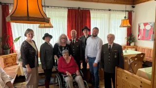 A group of people in a room with a woman in a wheelchair, dressed in formal attire, standing around her with some wearing hats. There are curtains, lamps, and a table with glasses.