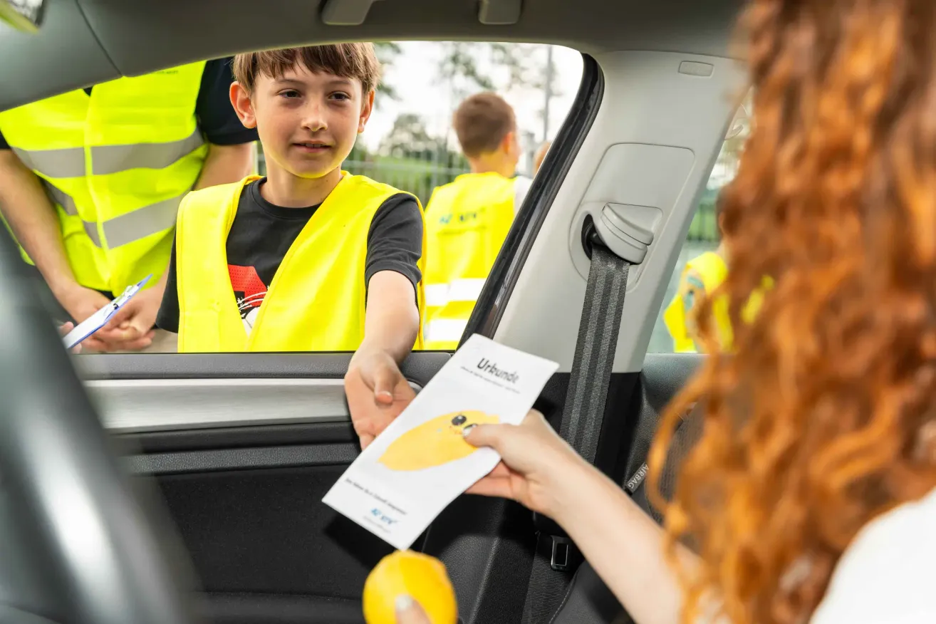 A young boy in a yellow vest hands a piece of paper to a woman, likely participating in an event. Other people in yellow vests are in the background.