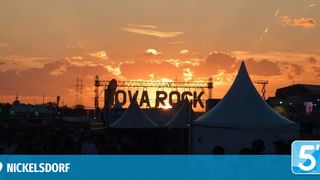 Ein Festival bei Sonnenuntergang mit Menschen in Zelten und einer Bühne. Die Sonne geht hinter der Bühne unter.