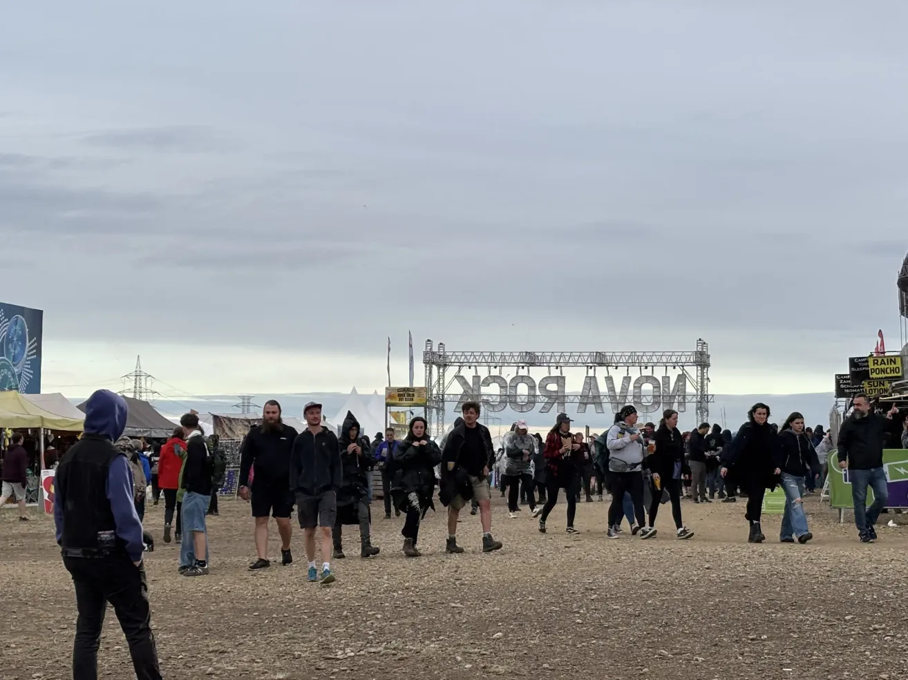 Viele Menschen laufen auf einem offenen Gelände unter einem bewölkten Himmel, mit einer großen Bühnenstruktur im Hintergrund.