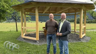 Zwei Männer stehen vor einem hölzernen Pavillon auf einer Wiese mit einem Berg im Hintergrund.